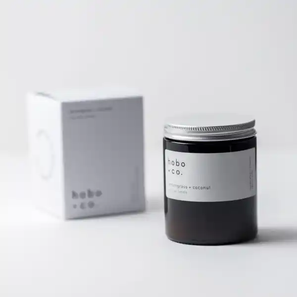 Eine Duftkerze von hobo + co. mit Zitronengras und Kokosnuss, neben der Verpackung auf einem weißen Hintergrund.