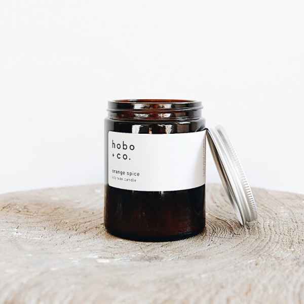Braunes Glas mit weißem Etikett und silbernem Deckel, enthält eine Duftkerze von hobo + co. auf einem Holzuntergrund.
